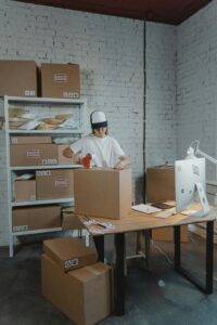 A warehouse worker in uniform packaging boxes for delivery in an organized storage area.
